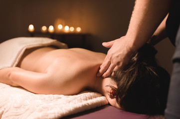 Men's hands make a therapeutic neck massage for a girl lying on a massage couch in a massage spa with dark lighting. Close-up. Dark Key