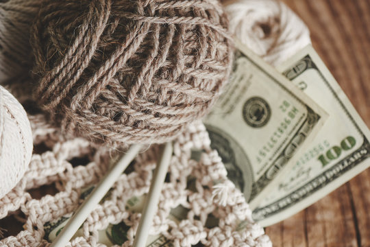Profitable hobby. Earnings on needlework. Balls of natural color yarn, knitting needles and money on a wooden table.