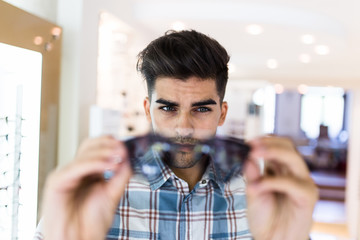 Handsome young man choosing eyeglasses frame in optical store.