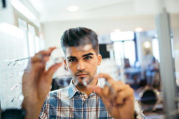 Handsome young man choosing eyeglasses frame in optical store.