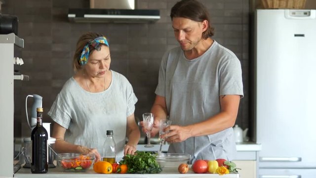 Young man and a mature woman are cooking dinner in the kitchen. He puts glasses on the table. On the table are ripe vegetables and a bottle of wine. Couple with a difference in age