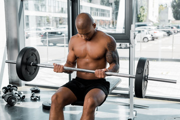 Fototapeta premium athletic bare-chested african american sportsman sitting on bench and lifting barbell in gym
