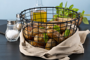 Baked potatoes in a black basket. Basil, olive oil, salt, pepper. The background is light blue. Copy space. Horizontal shot.