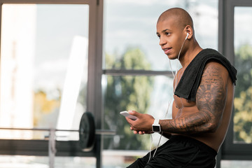 smiling young muscular bare-chested african american man in earphones sitting and using smartphone in gym