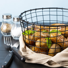 Baked potatoes in a black basket. Knife and fork. Basil, salt, pepper. The background is light blue. Copy space. A square shot.