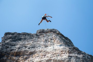 From below shot of climber secured with rope jumping from top of mountain under blue sky 