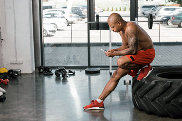 Fototapeta na wymiar side view of shirtless african american athlete resting and using smartphone on tire at gym