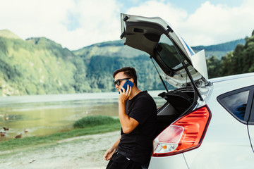 Young travel man in sunglasses sitting in car trunk talk on phone on nature lake landscape on background. Summer travel.