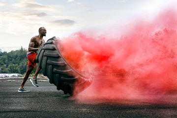 Handsome african american muscular man flipping burning big tire outdoor with smoke