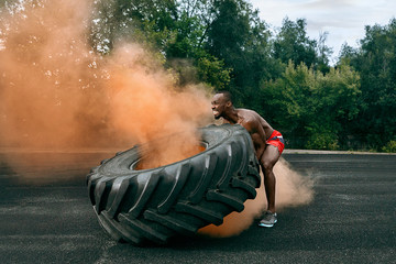 Obraz premium Handsome african american muscular man flipping burning big tire outdoor with smoke