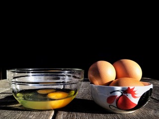 Eggs yolk in glass bowl and fresh chicken eggs in ceramic cup on wooden table floor with sunlight on surface in black background, close up shot with copy space