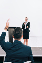 selective focus of attractive speaker talking into microphone during seminar in conference hall, participant raising hand