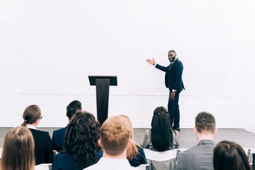 smiling african american lecturer talking to audience during seminar in conference hall and pointing on screen