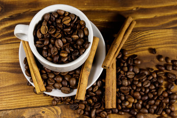 Coffee beans in white cup and cinnamon sticks on wooden table. Top view