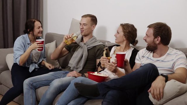 Friends are gathering together and having fun at the living room with loft interior. Male and female company, The girl with a big red bowl with popcorn, everyone drinking beer or soda, laughing