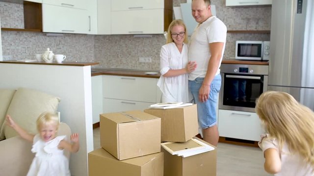 Young happy family moving to new apartment. Two little girls running into new home with parents at background cardboard boxes, happy couple with children moving in new house. Relocating, housewarming