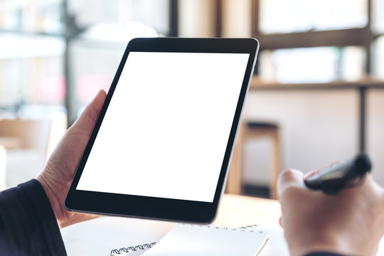Mockup image of hands holding and using black tablet pc with blank white desktop screen while writing on notebooks in office