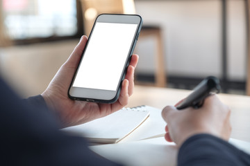 Mockup image of hands holding white mobile phone with blank white screen while writing on notebook in office