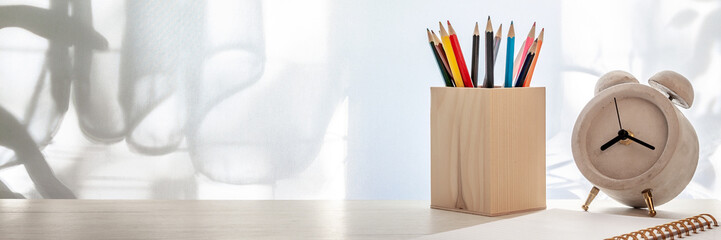 Panoramic photo of a white wooden desk with colored pencils in a wooden toolbox, open notebook and alarm clock with concrete on the background of sun-lit curtains. Copy space