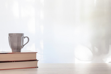 Photo of a white desk with thick books and a gray cup of hot tea on a background of sun-lit curtains. Copy space