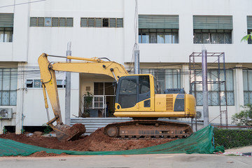 Excavator is digging soil on the ground behind the building in construction area.