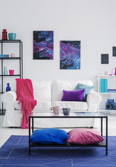 Pillows under table on blue carpet in front of white couch in flat interior with posters. Real photo