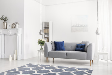 Patterned blue carpet in front of grey sofa in white apartment interior with flowers and lamps. Real photo