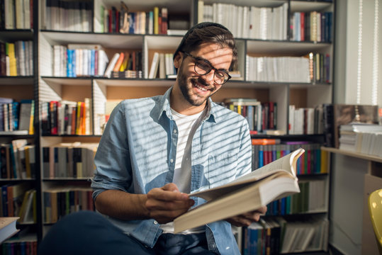 Happy male student sitting in library and reading book. In background bookshelf.