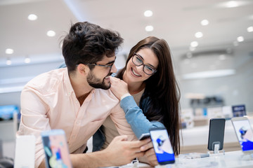 Naklejka premium Man and woman talking about which smart phone to buy. Man holding in hands smart phone while woman leaning on him.