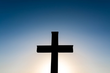 Simple oak wood catholic cross silhouette at sunset, dramatic dark blue sky with the glowing sun.