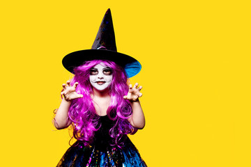 Small girl in Halloween witch dress and hat scaring and making faces isolated on yellow background.