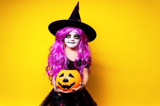 Small girl in Halloween witch dress and hat scaring and making faces isolated on yellow background. Trick or treat.