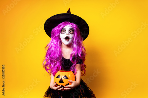 Small girl in Halloween witch dress and hat scaring and making faces isolated on yellow background. Trick or treat.