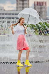 Young pretty girl with two braids in yellow boots and with transparent umbrella stands near fountain.