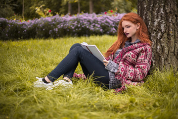 Fototapeta premium Redhead young concentrated woman with freckles and dark lipstick reading book outdoors in park sit under the big tree on grass with flowers on the background.