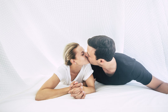 Real love for caucasian couple under the sheets at home in the bed and bedroom. bright white image and true partnership with man and woman together taking hands and kissing