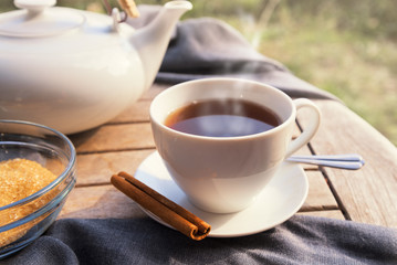 black tea in a cup, a white teapot, brown sugar and cinnamon on a wooden garden table