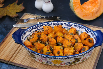 Baked pumpkin slices with garlic and seeds in a ceramic form on a dark background. On the table are jars of salt and pepper and a piece of pumpkin