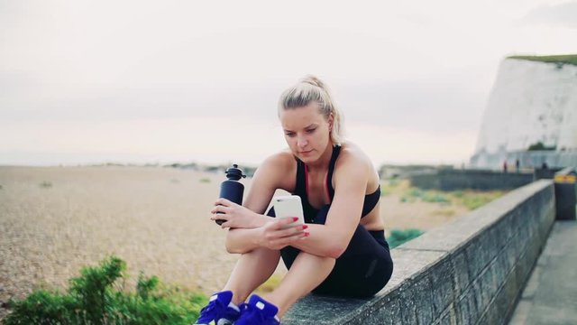Young sporty woman with smartphone sitting outside, texting. Slow motion.