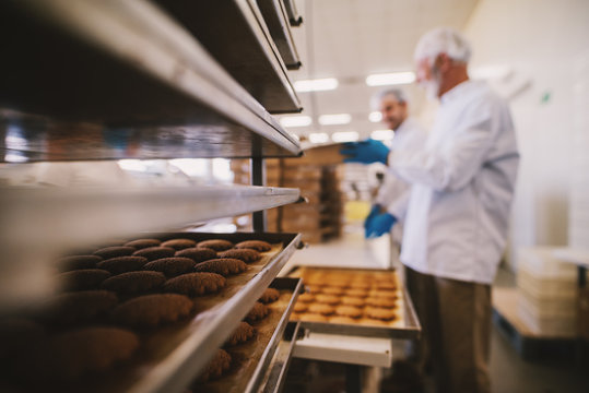 Close up of tray full of fresh baked cookies in food factory. Blurred picture of two male employees in sterile clothes in background.