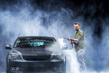 A man with a beard or car washer washes a gray car with a high-pressure washer at night in a shop...
