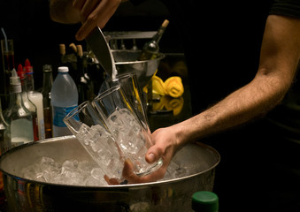 Bartender`s hand putting ice into the cocktail glass on the grey blurred background on bar counter  Barman holds three glasses and  putting broken ice in the glasses