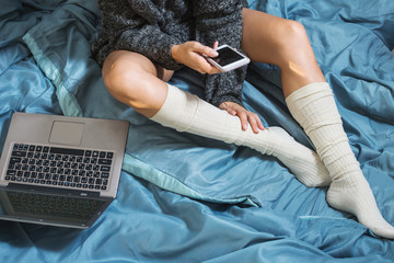 A young girl with sexy legs in a warm sweater lying on the bed with smartphone and laptop. Work at home