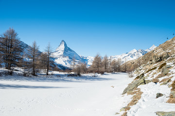 Fototapeta premium View on winter forest with pine trees, snowy Matterhorn, frozen lake Grindjisee, mountains and blue sky with white clouds, Switzerland.