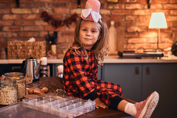 Cute little girl in rabbit makeup hat and pajamas sitting on the table with scattered food in loft style kitchen at morning.