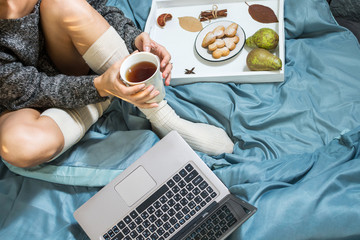 Young woman drinking tea at home in her bed and checking her laptop