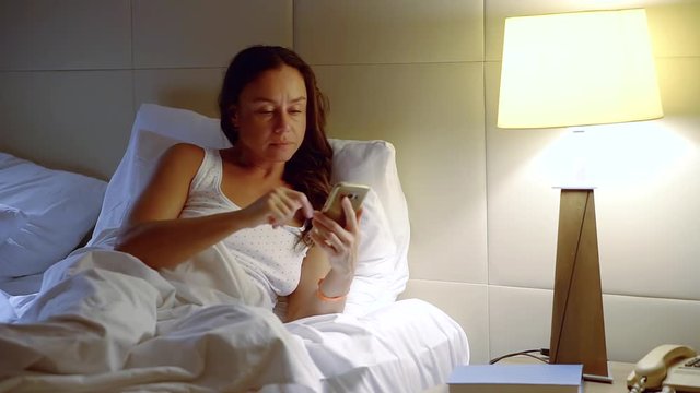 brunette woman is using smartphone lying in a bed, switching off a table lamp
