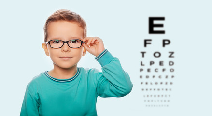 Little boy adjusting his new eyeglasses, on the eye chart background. Vision correction for children