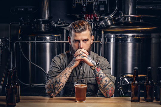 Portrait of a pensive tattooed hipster male with stylish beard and hair in the shirt in indie brewery.  