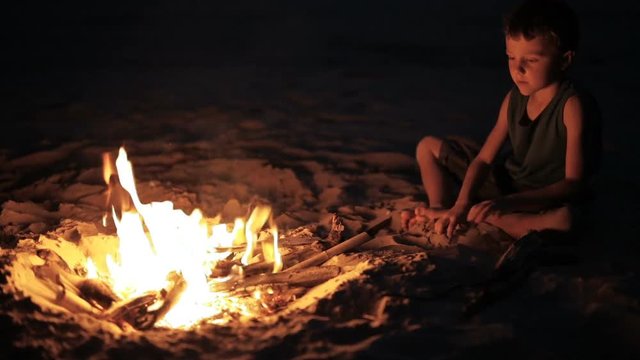 Happy little boy sitting near the bonfire on the beach at the sunset time